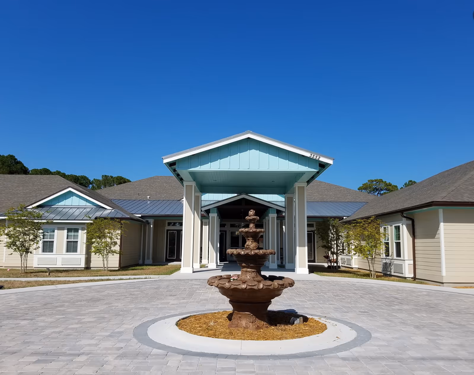 Front entrance of a single-story building with a covered portico and a decorative fountain in a circular drive under a clear blue sky.