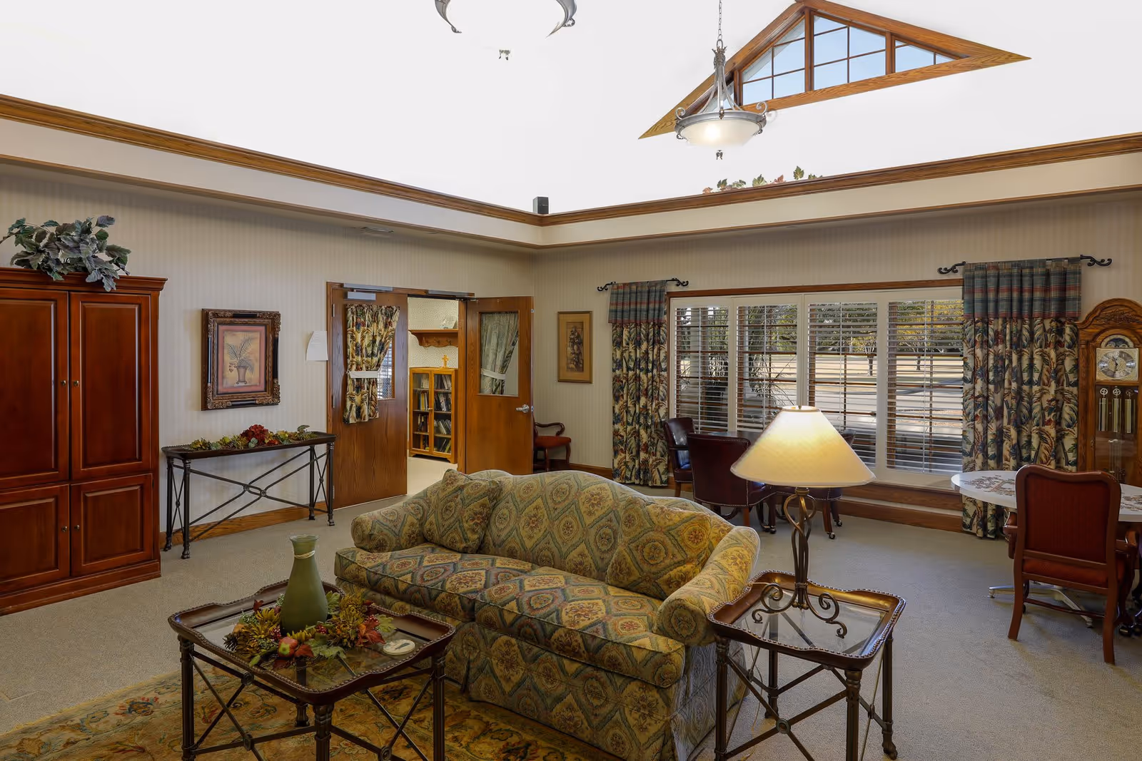 A cozy senior living facility common area with a patterned sofa, glass-top side tables, a table lamp, and a coffee table decorated with a green vase and autumn-themed centerpiece. The room has large windows with floral curtains, a grandfather clock, and a wooden cabinet. The ceiling features a skylight with wooden beams and hanging light fixtures.