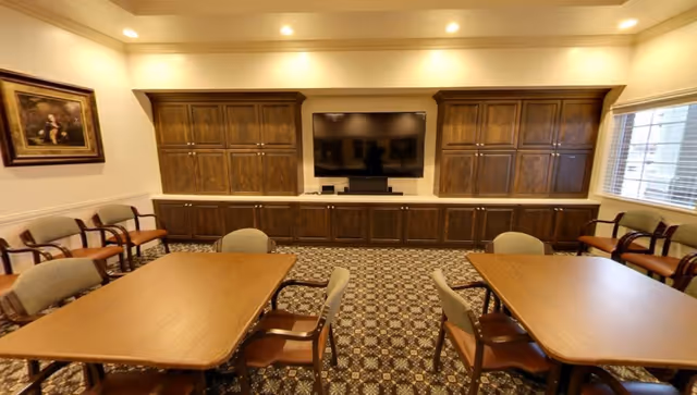 A senior living common room with tables and chairs facing a large TV set within wooden built-in cabinets.