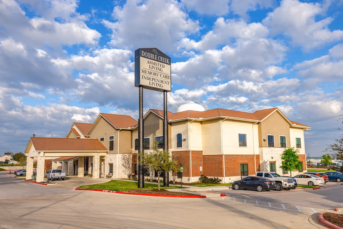 Exterior view of Double Creek Assisted Living and Memory Care facility showing a two-story building with beige and brick facade, a covered entrance, several parked cars, and a tall sign displaying the facility's name and services under a partly cloudy blue sky.