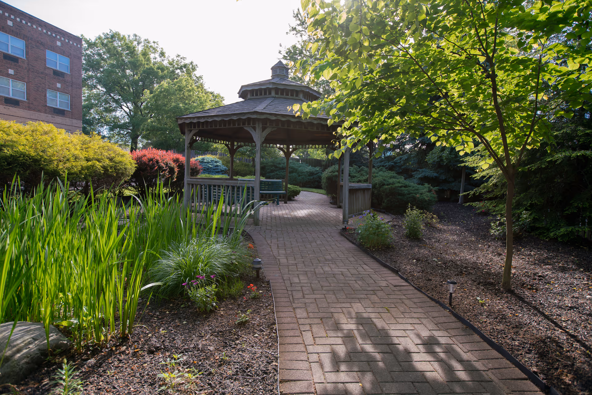 A brick-paved garden path leads to a wooden gazebo surrounded by trees, shrubs, and a nearby building.