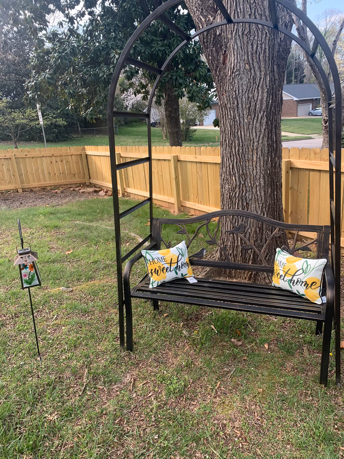 A black metal garden bench with decorative bird and branch design, placed under a large tree in a grassy backyard. The bench has two cushions with yellow flowers and the words 'Home sweet home' printed on them. A wooden fence encloses the yard, and a decorative lantern on a stake is positioned to the left of the bench.