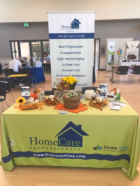 Promotional table with HomeCare Professionals banners and fall decorations at an expo, with attendees in background.