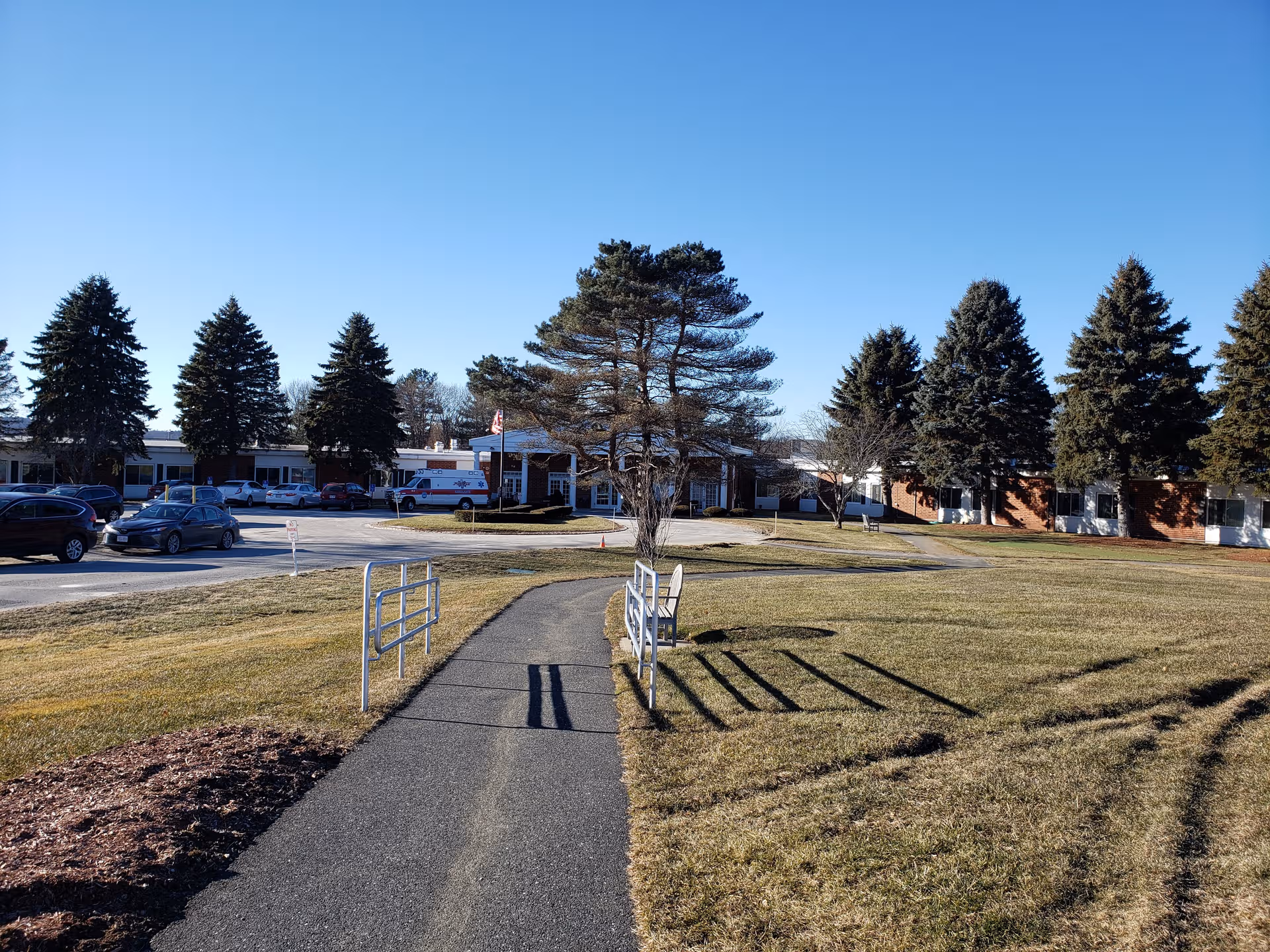 Exterior view of Life Care Center of Nashoba Valley with a walkway, front entrance, and cars.