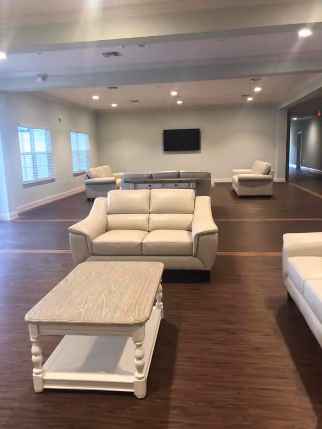 A spacious living room with multiple beige leather sofas arranged around a wooden coffee table. The room has wooden flooring, white walls, and a flat-screen TV mounted on the far wall. Large windows on the left side allow natural light to enter the room.