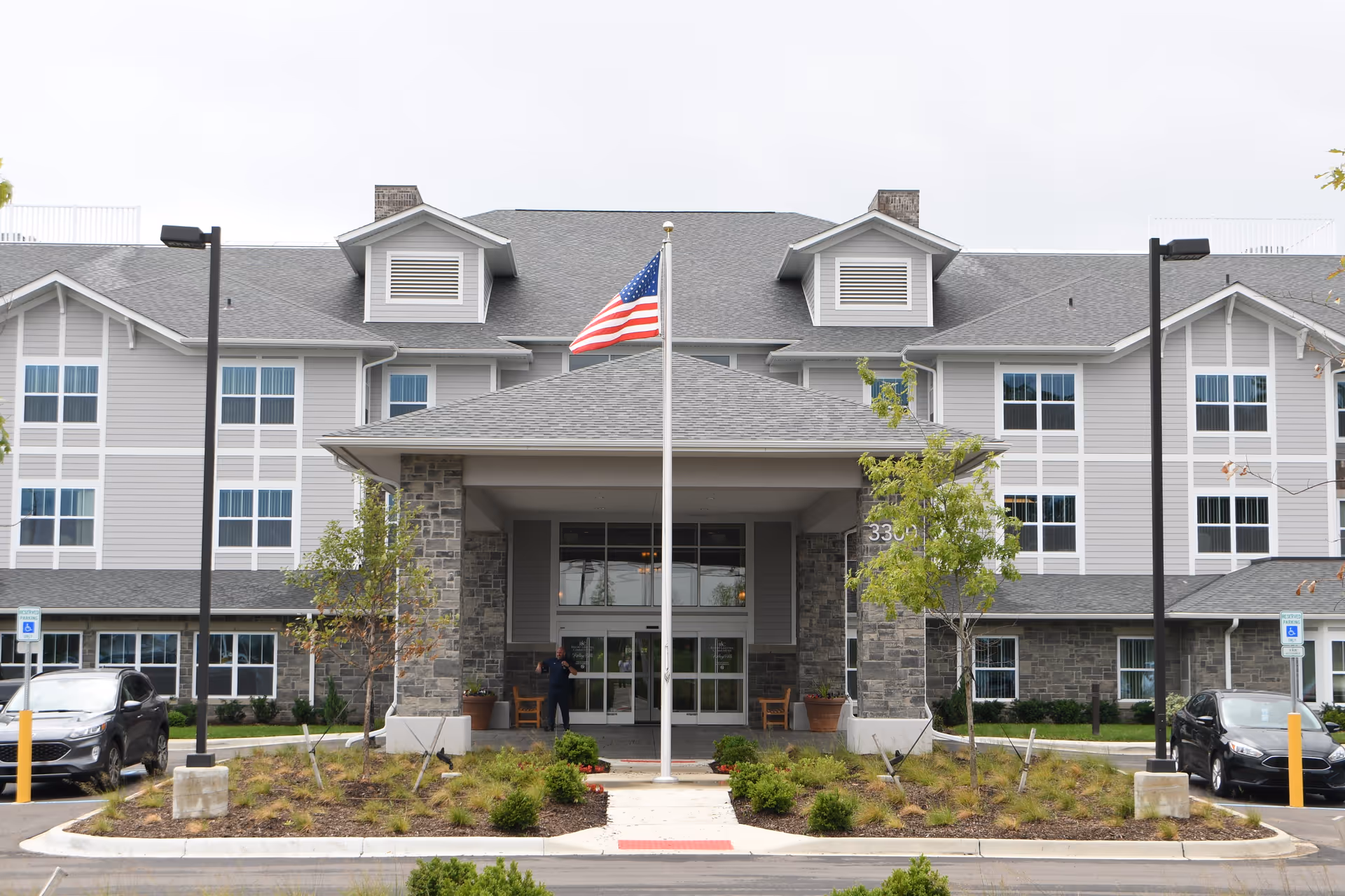 Front exterior view of Rolling Hills Retirement Community building with a covered entrance, an American flag on a flagpole in front, two parked cars, and landscaped greenery.