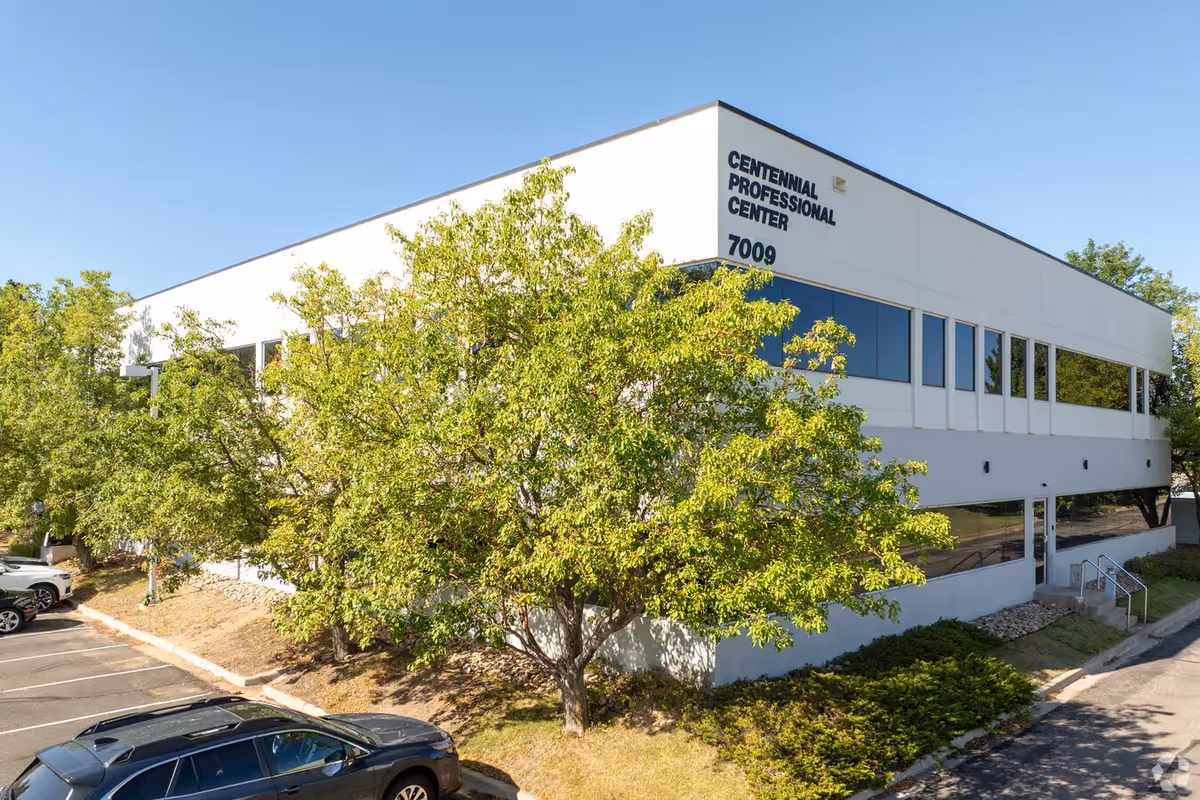 White modern two-story building with large windows, trees, and a parking lot.