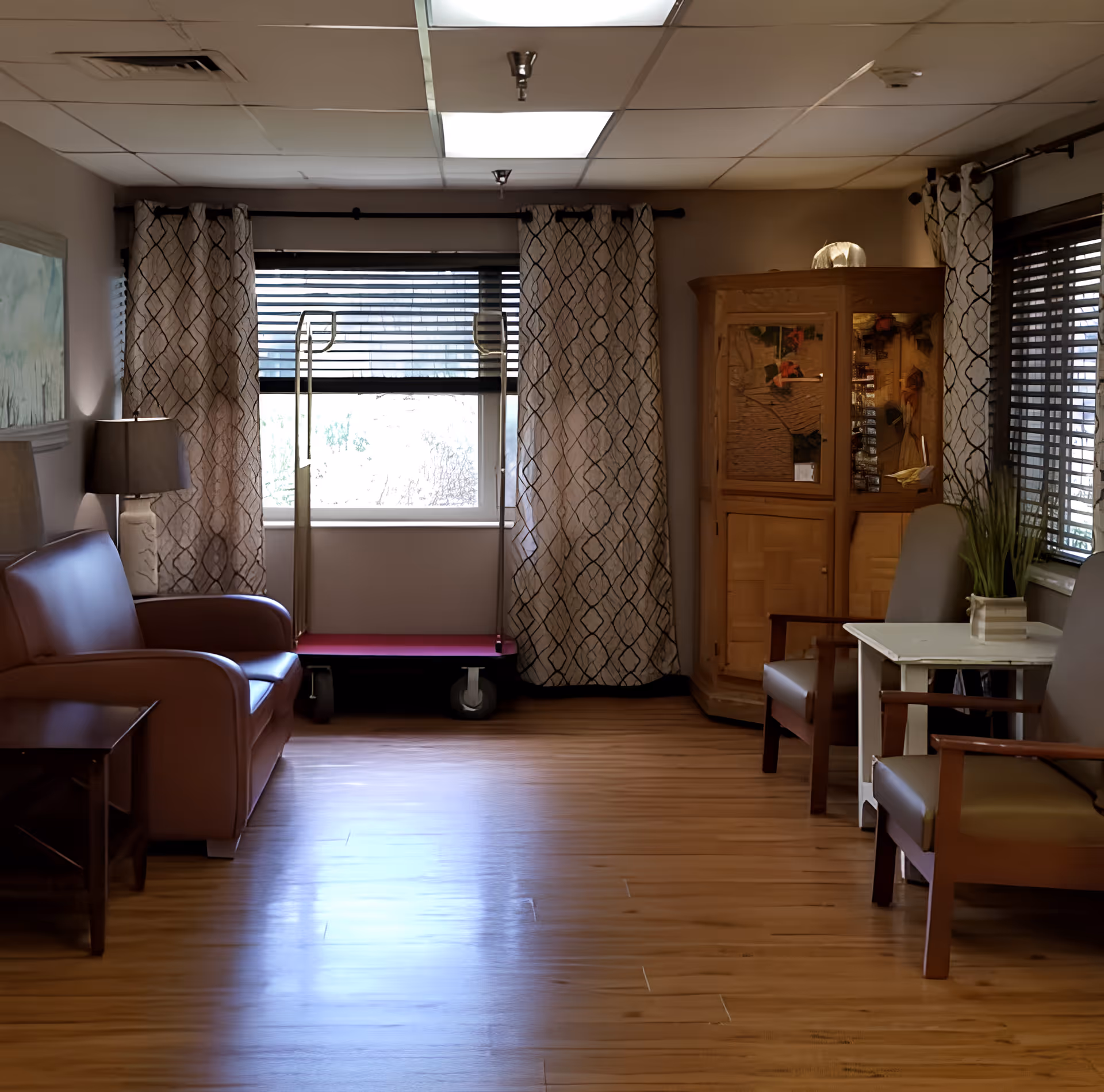 A cozy interior room with wooden flooring, featuring a brown leather sofa on the left, a wooden side table with a lamp, two armchairs with a small white table between them on the right, a wooden cabinet with glass doors in the corner, and windows with patterned curtains and blinds.