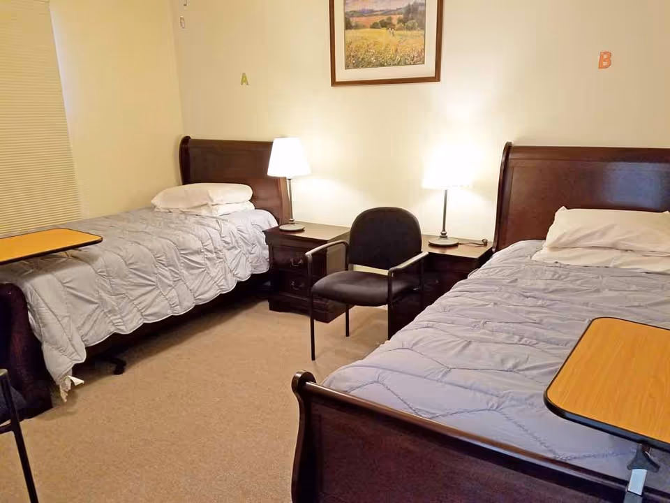 A bedroom with two single beds, each with a light gray comforter and white pillows. Between the beds are two dark wooden nightstands with lamps on each. A black chair is placed between the nightstands. There is a framed landscape painting on the wall above the nightstands. Both beds have overbed tables positioned at the foot. The room has beige carpet and light-colored walls.