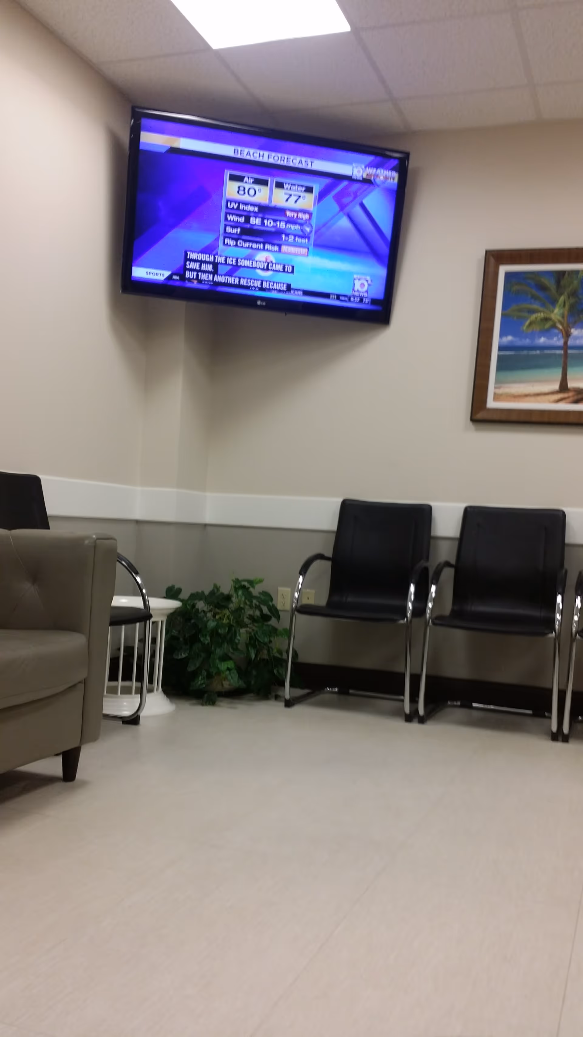 Indoor waiting area with wall-mounted TV, black chairs, plant, and framed beach painting.