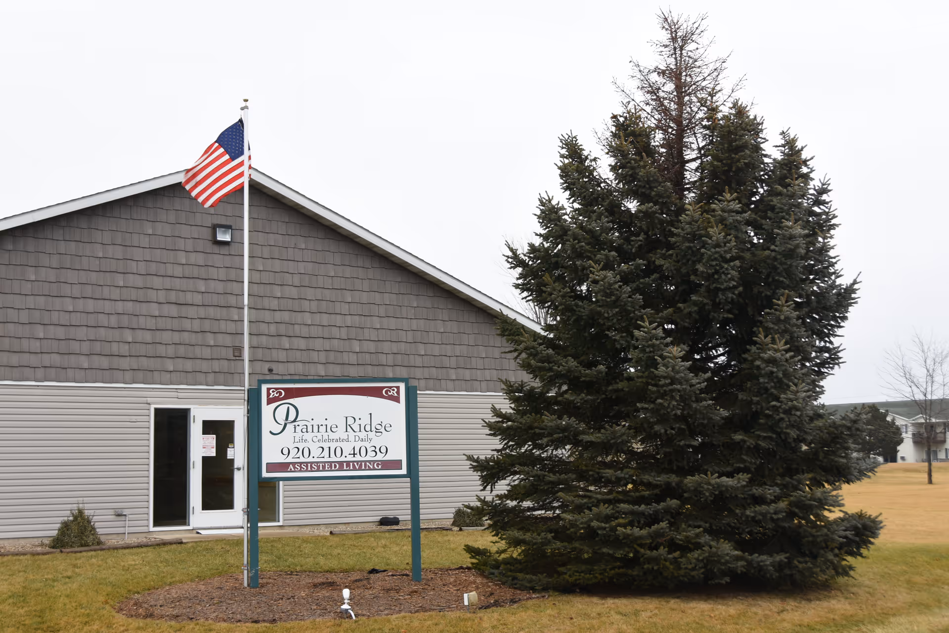 Prairie Ridge Assisted Living building with sign, flagpole, and a large evergreen tree in front