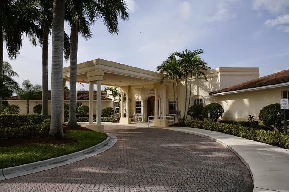 Cream-colored senior living facility with a covered entrance, palm trees, and brick driveway.