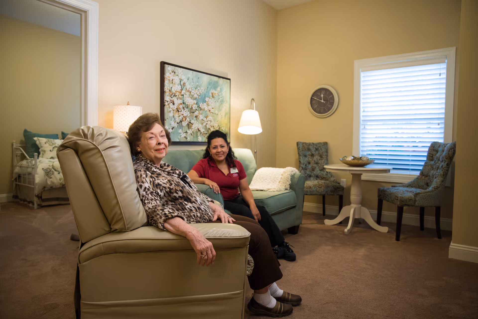 An elderly woman sitting in a beige recliner chair in a cozy living room area with a younger woman sitting on a green couch behind her. The room has beige walls, a painting of white flowers, a floor lamp, a round table with two patterned chairs near a window with blinds, and a clock on the wall. A bedroom with a bed and pillows is visible through an open doorway.