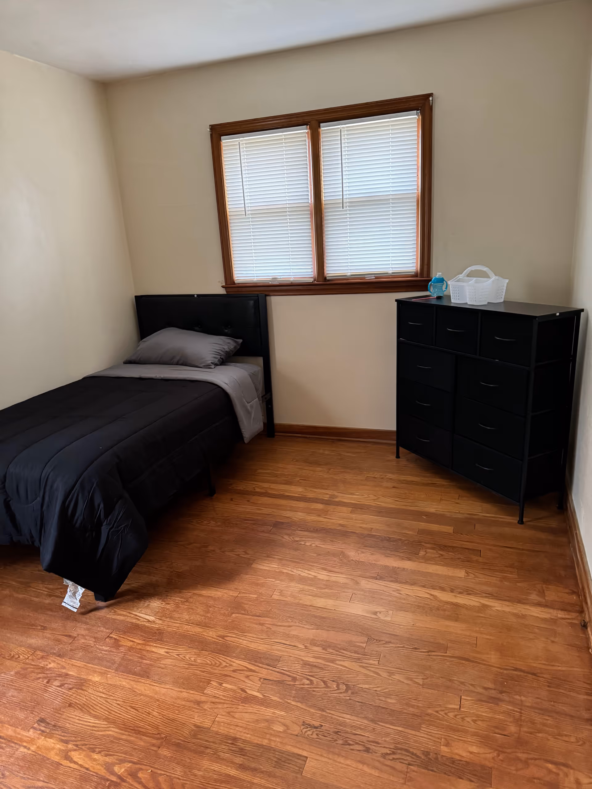 Small bedroom with a black bed, dresser, window blinds, and wooden floor.