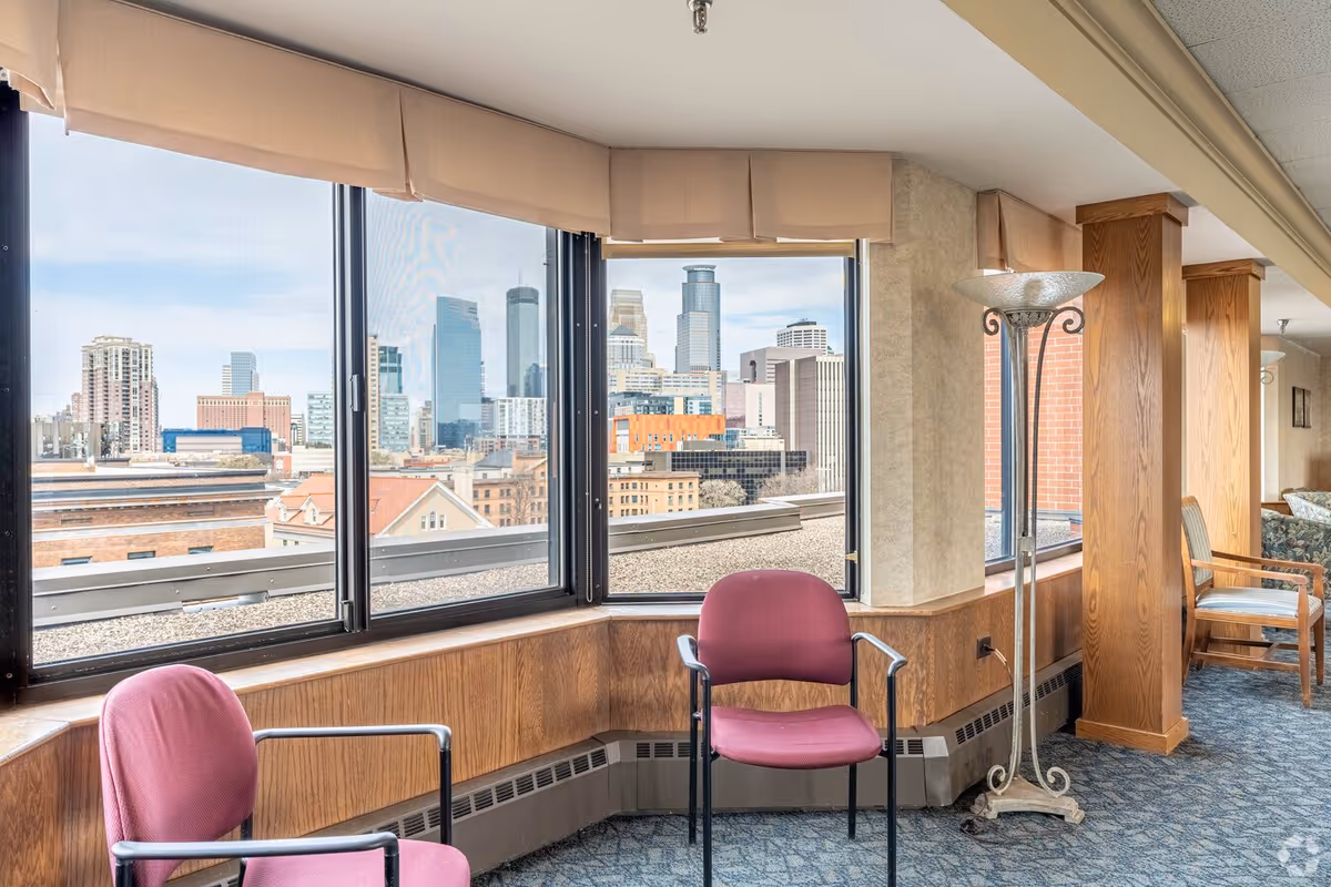 Interior senior living lounge with pink chairs and large windows overlooking cityscape.