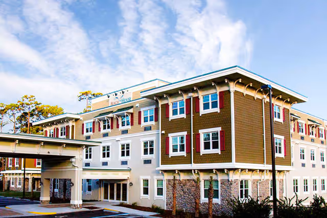 Front exterior of a three-story assisted living building with a covered porte-cochère, stone base, and rows of windows with red shutters under a blue sky.
