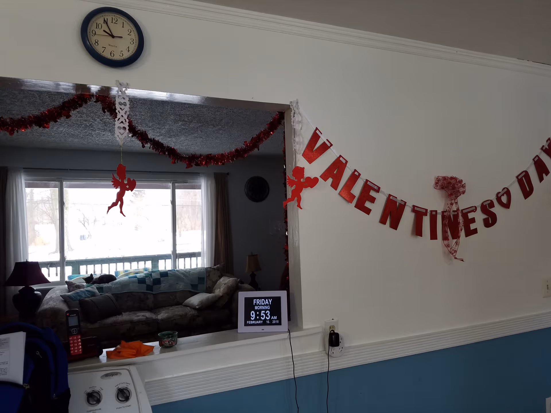 Interior view of a senior living facility decorated for Valentine's Day with red garlands and a banner spelling VALENTINES DAY. A clock on the wall shows the time as 9:53. Through a pass-through window, a living room with a couch, lamps, and a large window with curtains is visible. A digital calendar displays the date as Friday, February 16, 2018.