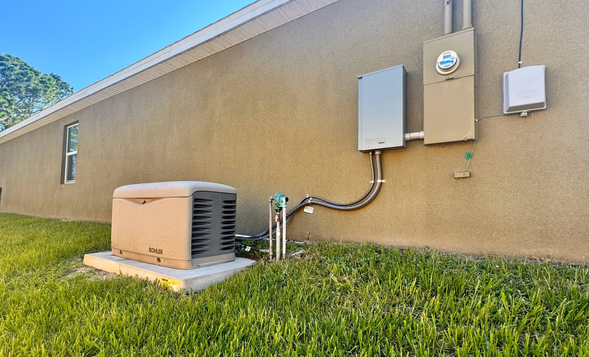 Exterior side view of a building wall with a Kohler generator placed on a concrete slab surrounded by grass. Electrical boxes and conduit pipes are mounted on the wall above the generator.