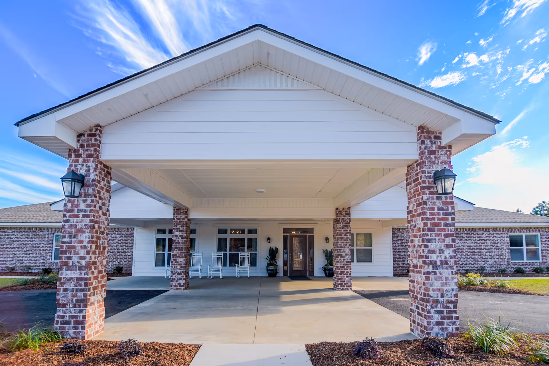 Front exterior view of a single-story brick building with a covered entrance supported by brick columns. The entrance area has rocking chairs and potted plants, under a white roof with a clear blue sky above.