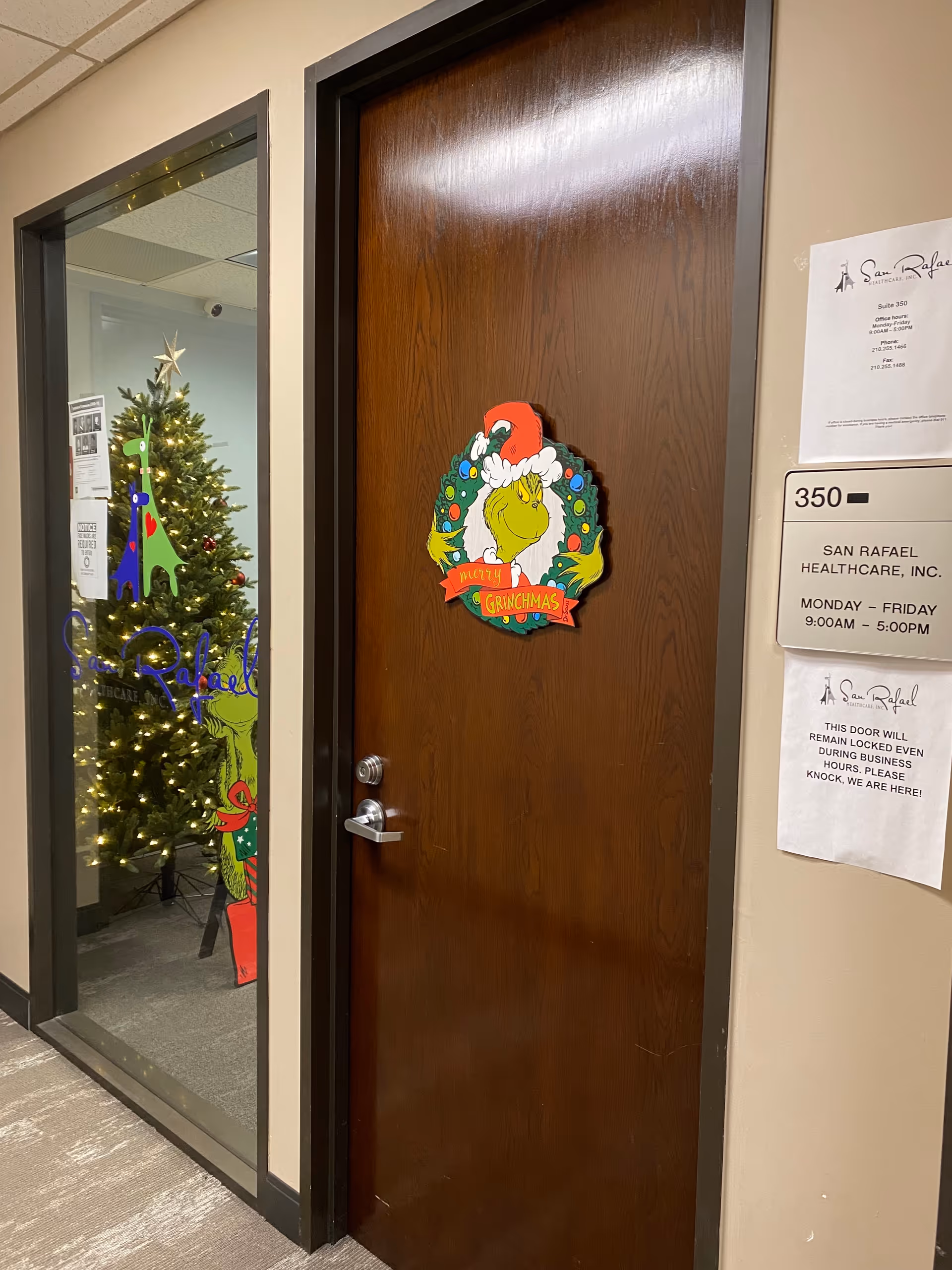 Wooden door with Grinchmas wreath; glass-panel hallway reveals a Christmas tree.