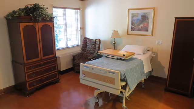 Hospice bedroom featuring a hospital bed, armoire, chair, lamp, and a window with blinds.