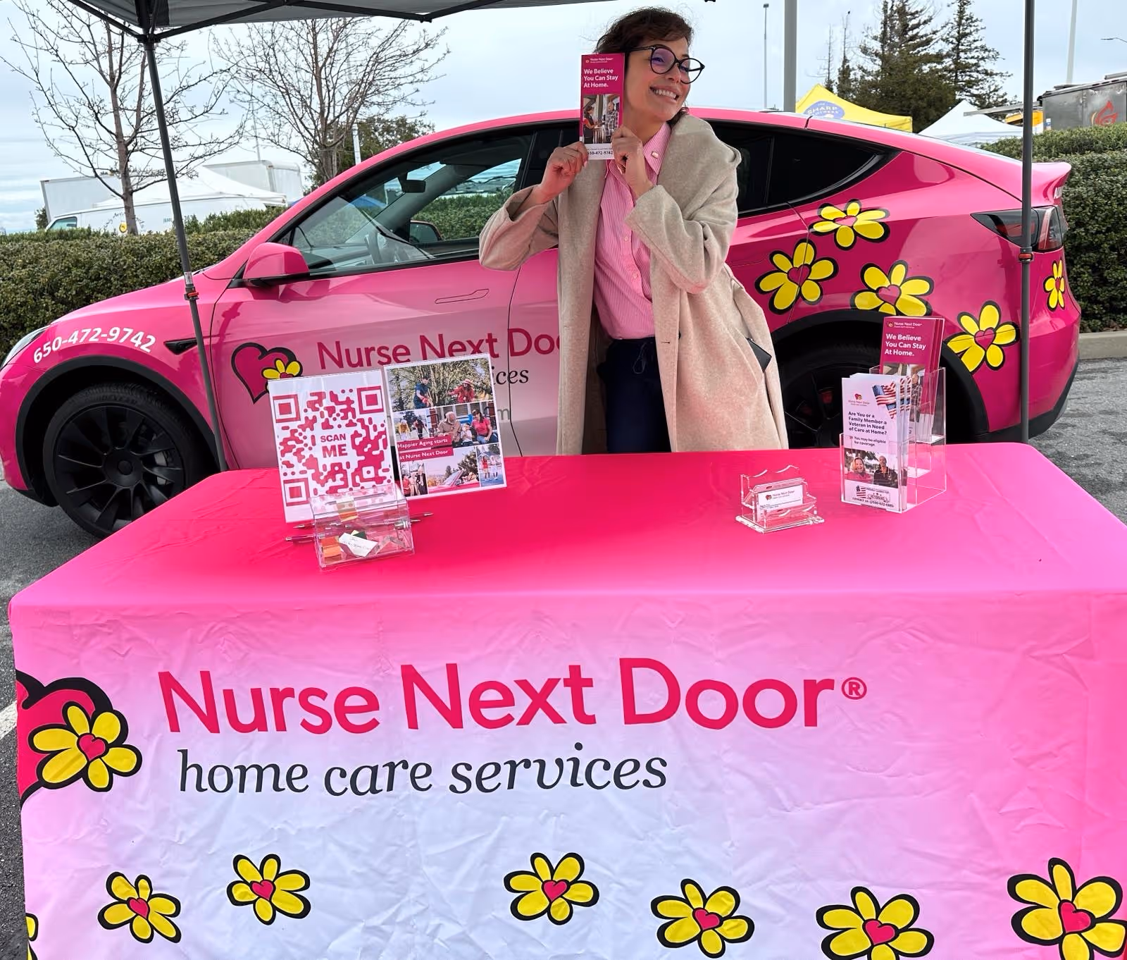 Staff member poses beside a pink Nurse Next Door car at an outdoor promotional booth.