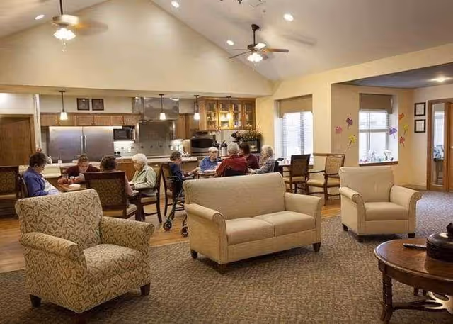 A spacious common area in a senior living facility with comfortable armchairs and sofas arranged on a carpeted floor. In the background, several elderly people are seated around tables near a kitchen area with stainless steel appliances and wooden cabinets. The room has high ceilings with ceiling fans and large windows letting in natural light.