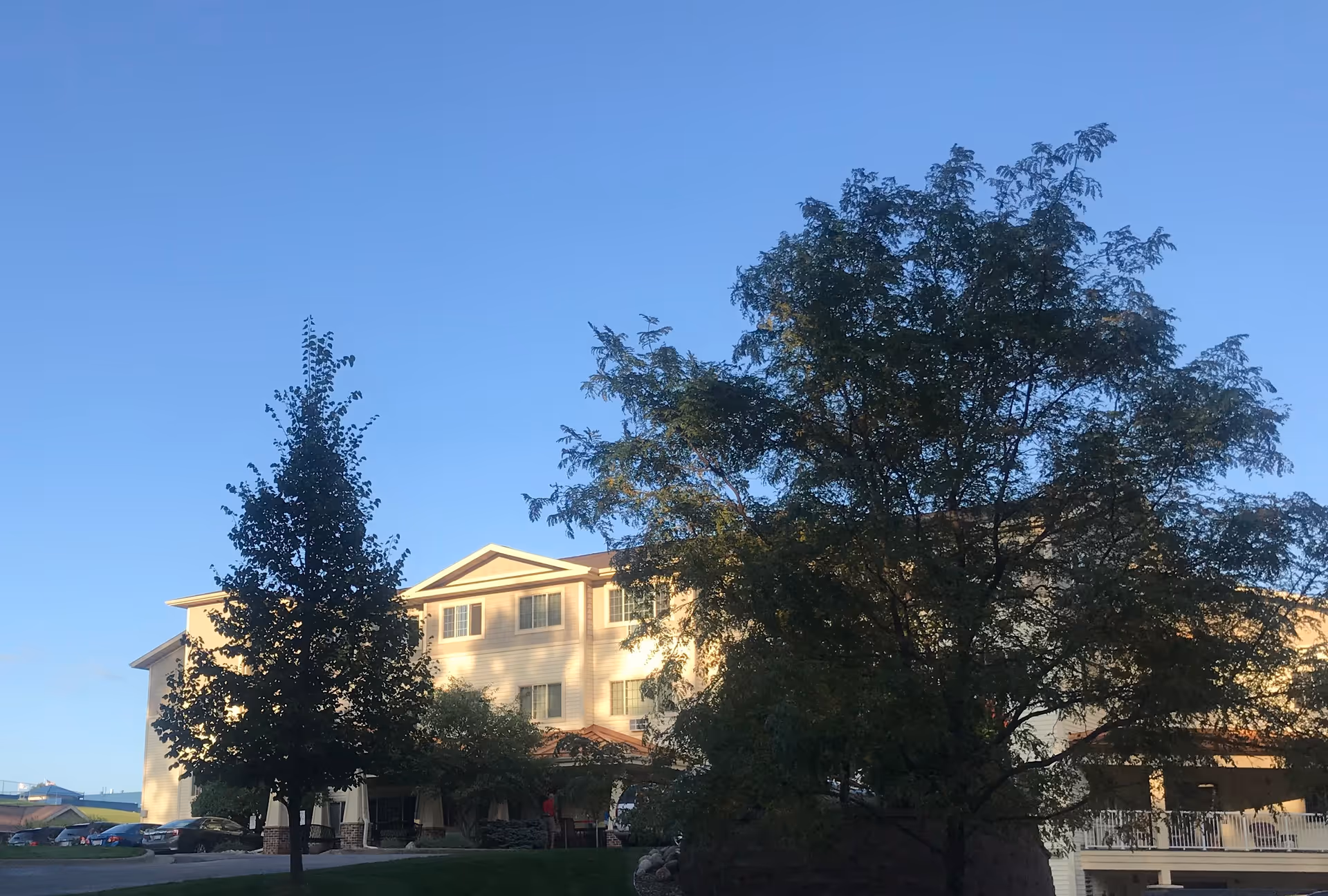 Exterior view of a multi-story assisted living facility building with several windows, surrounded by trees and a clear blue sky.