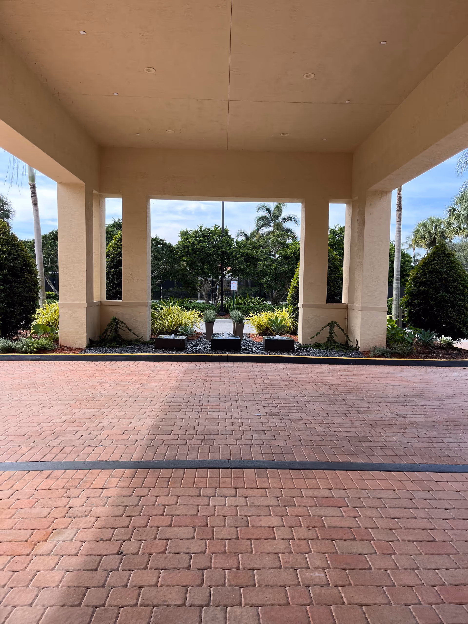 Covered entrance with beige columns and brick-paved driveway framed by landscaped plants.