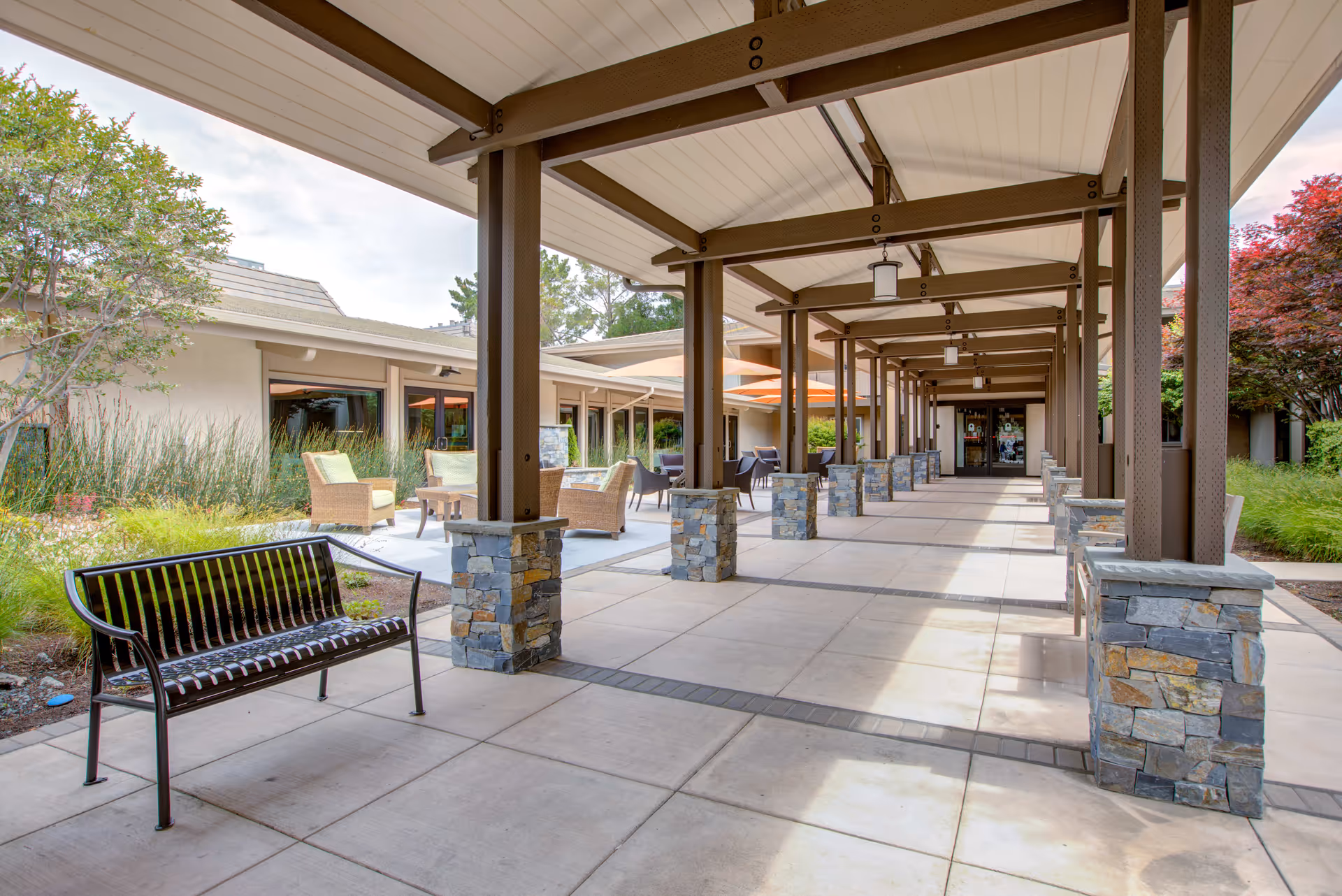 Covered outdoor walkway with stone pillars and brown beams at BridgePoint at Los Altos. There are benches and wicker chairs with tables arranged along the walkway, surrounded by greenery and trees.