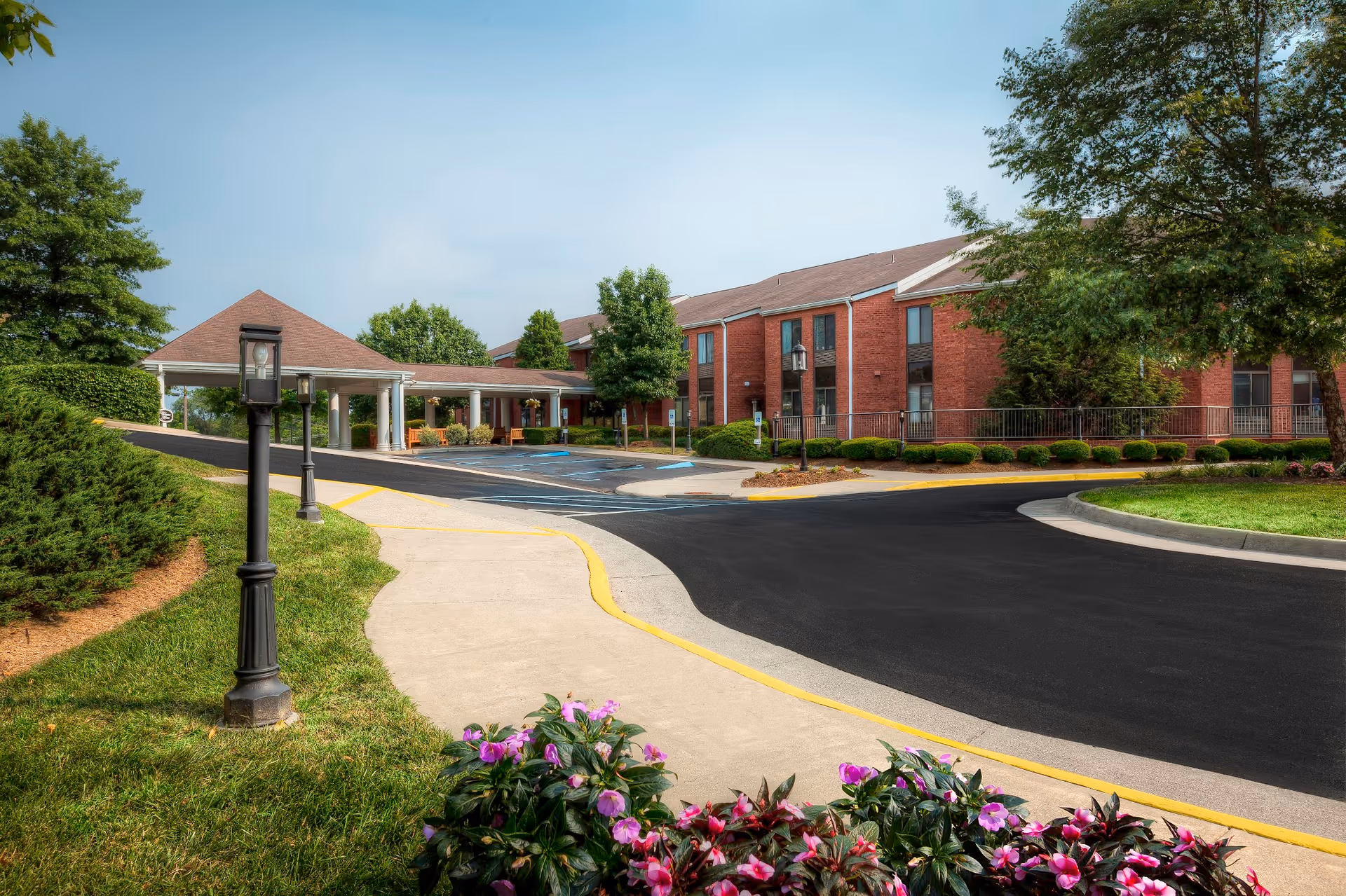 Exterior view of a senior living facility with a brick building, a covered entrance with white columns, a paved driveway, landscaped greenery, and flowering plants in the foreground under a clear sky.