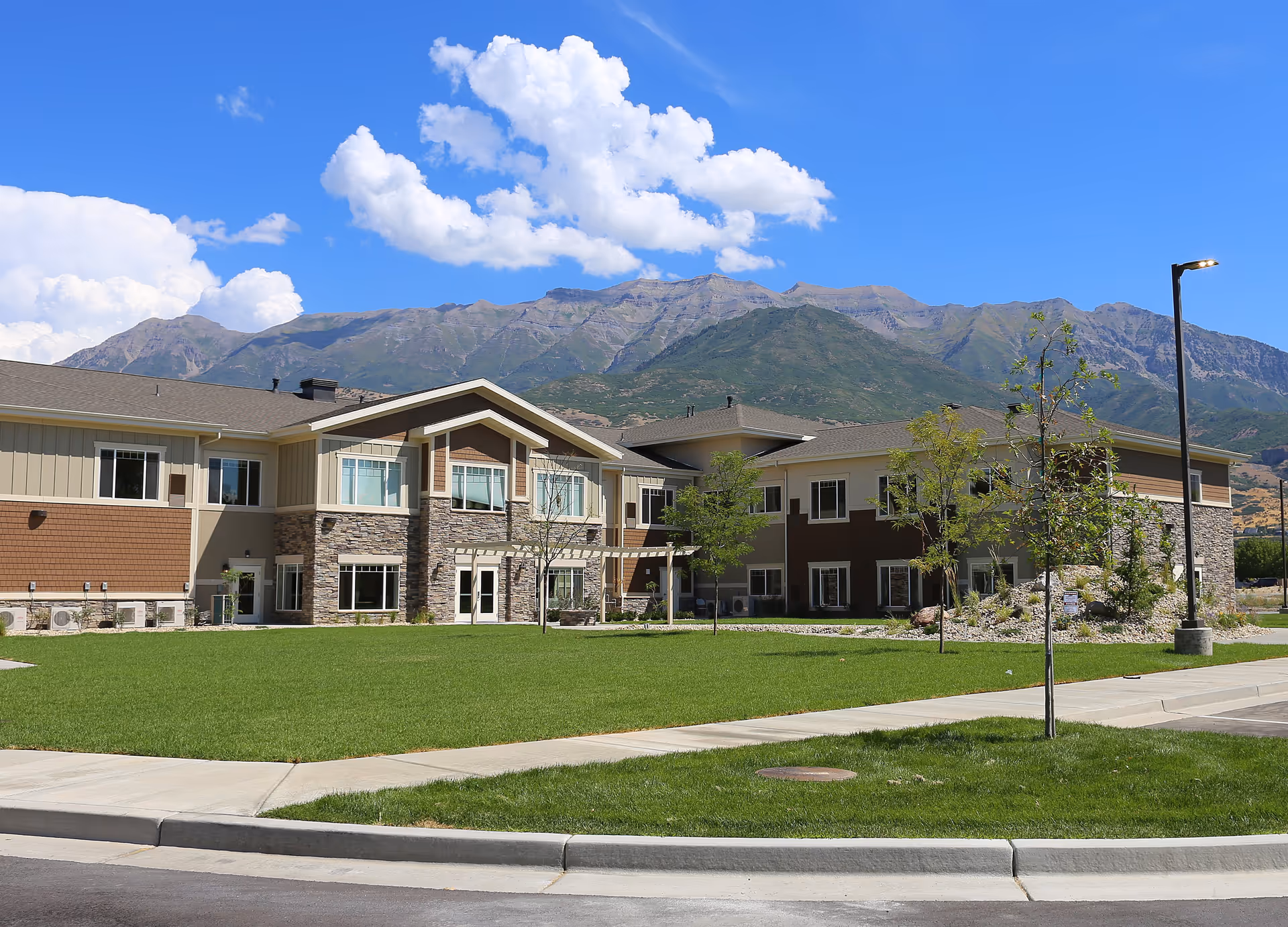 Exterior view of a two-story senior living facility building with a mix of stone and siding facade, surrounded by a well-maintained green lawn and young trees, with mountains and a blue sky with clouds in the background.