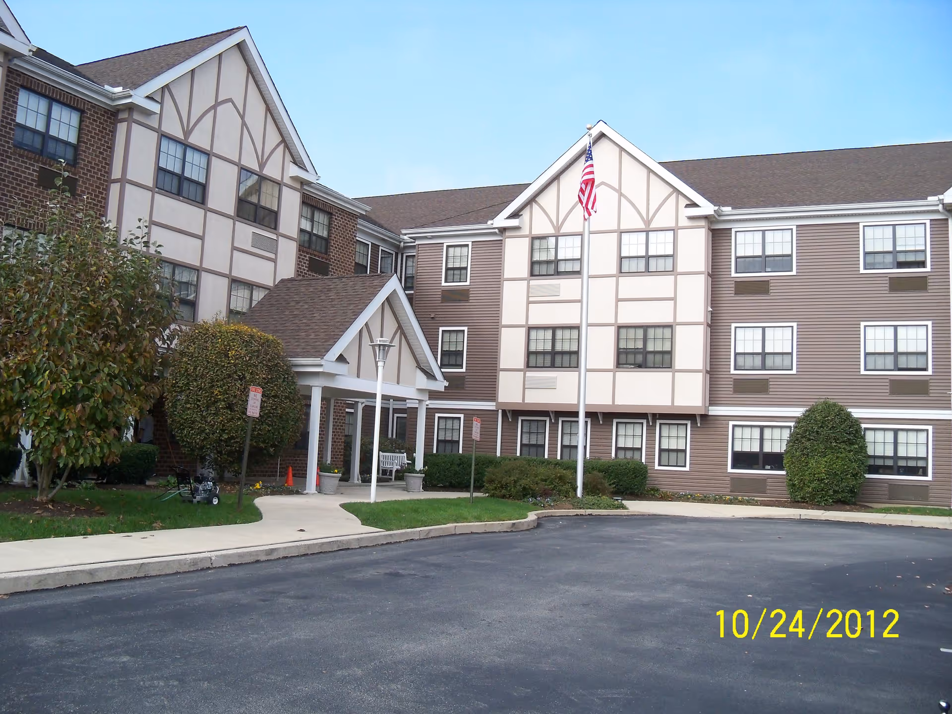 Front exterior of a multi-story senior living building with an entrance canopy, landscaping, and an American flag.