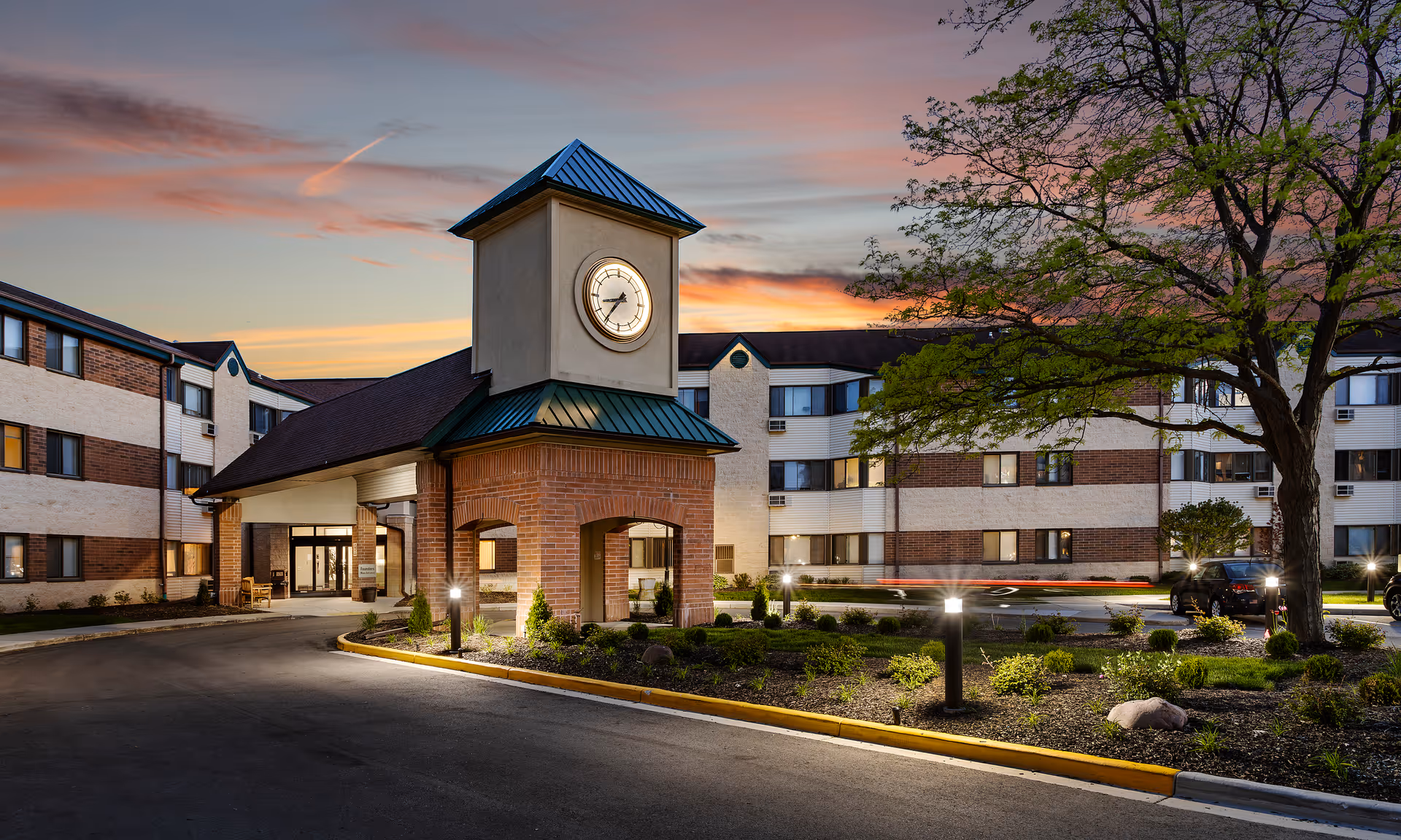 Front entrance of a senior living community with a clock tower, landscaped driveway and a multi-story brick-and-siding building at sunset.