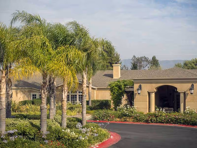 Exterior view of a single-story building with a beige facade, surrounded by palm trees and well-maintained landscaping including bushes and flowers, under a partly cloudy sky.