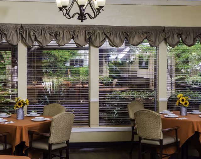 Dining room interior with round tables, orange cloths, sunflowers, and large windows.