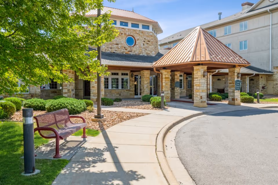 Covered porte-cochere entrance of a stone-faced senior living building with a bench, landscaped shrubs, and a curved driveway.