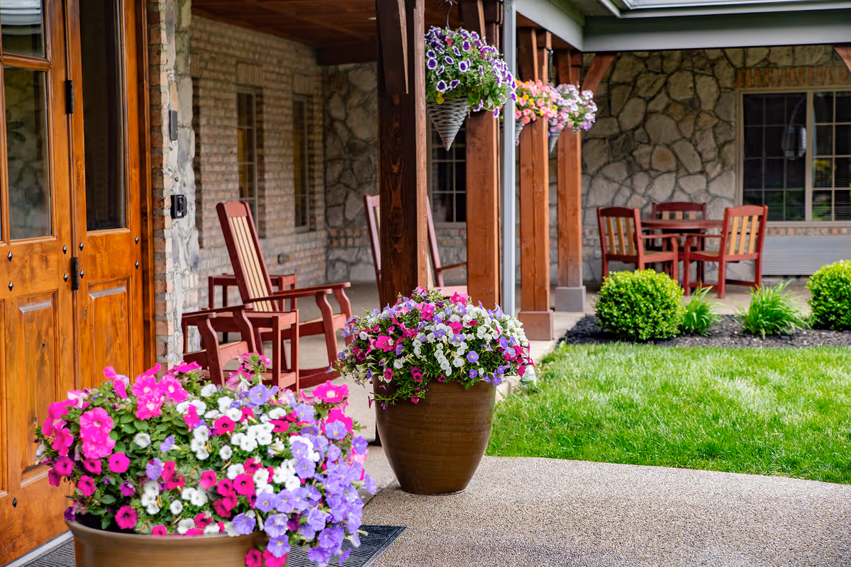 A covered outdoor patio with wooden chairs and colorful flower pots beside a stone building.