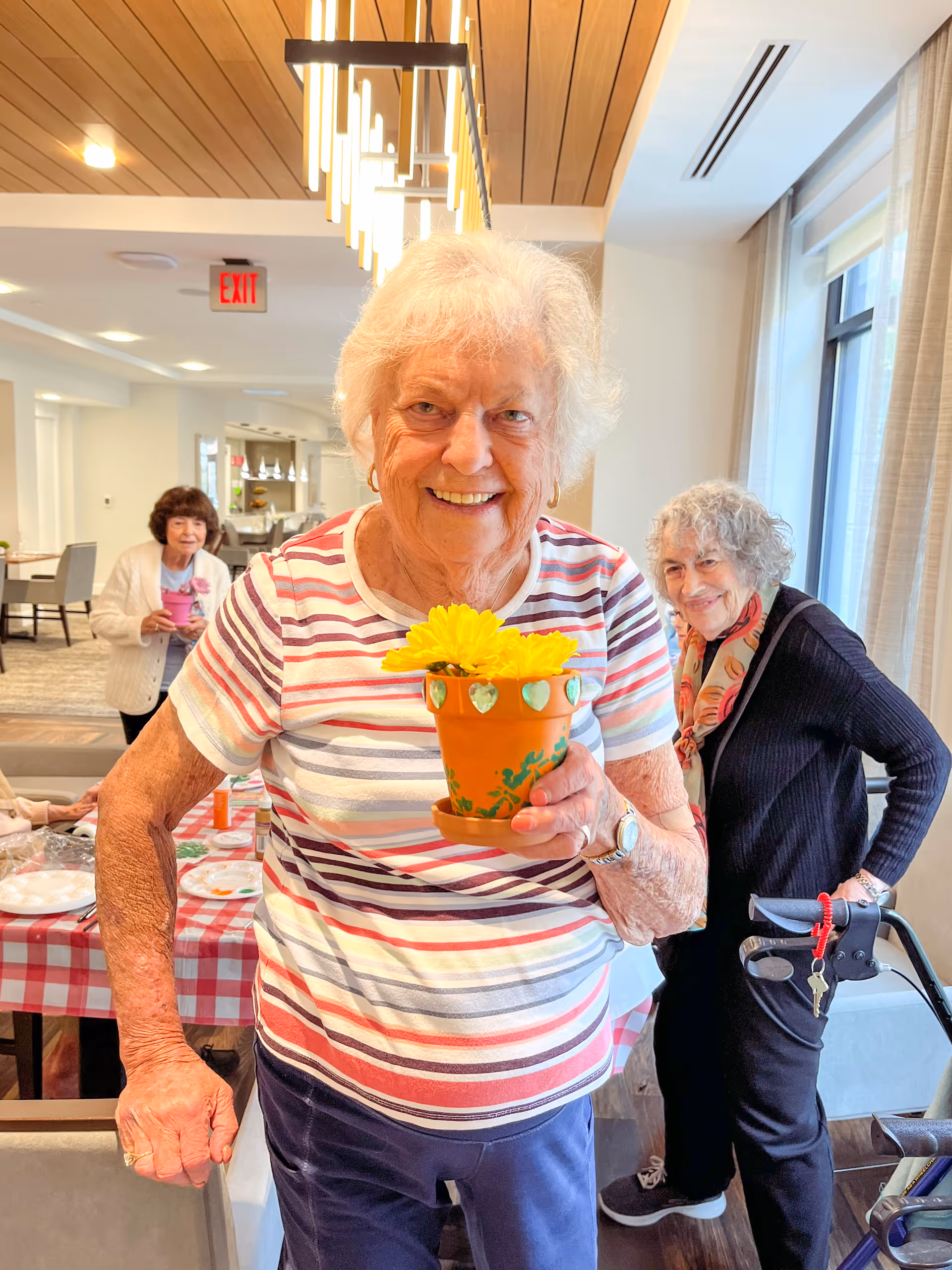 A smiling elderly woman in a striped shirt holds a small pot of yellow flowers in a bright communal dining/activity room with two other women in the background.