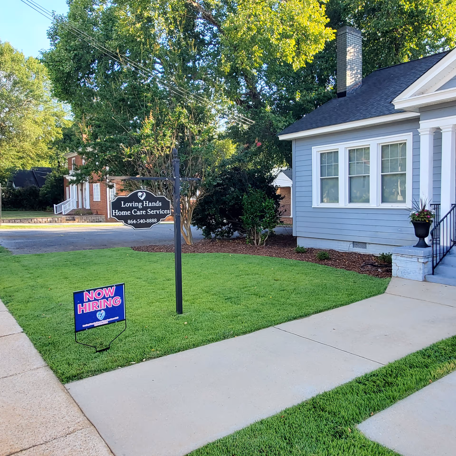 Front yard of Loving Hands Home Care Services with building sign and 'Now Hiring' sign.