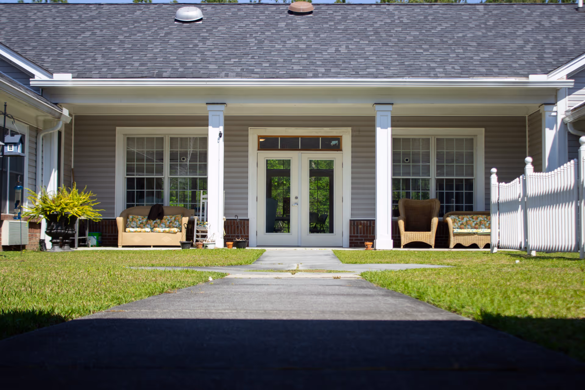 Front porch area of a building with a gray roof and beige siding, featuring two white columns flanking a set of double glass doors. There are two large windows on either side of the doors, with outdoor wicker furniture including chairs and a loveseat with floral cushions. A concrete pathway leads up to the porch, and there is a white picket fence on the right side.