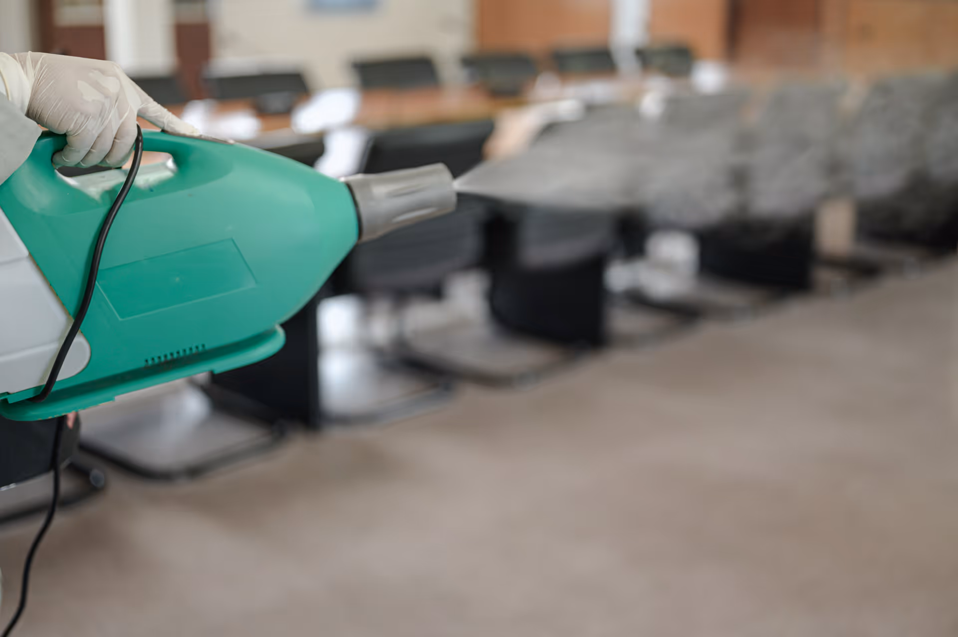 A gloved hand holds a green disinfecting sprayer, with a row of chairs in the background.