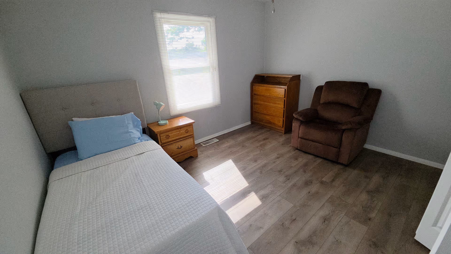 A tidy bedroom with a single bed, nightstand, window, dresser, and a brown recliner.