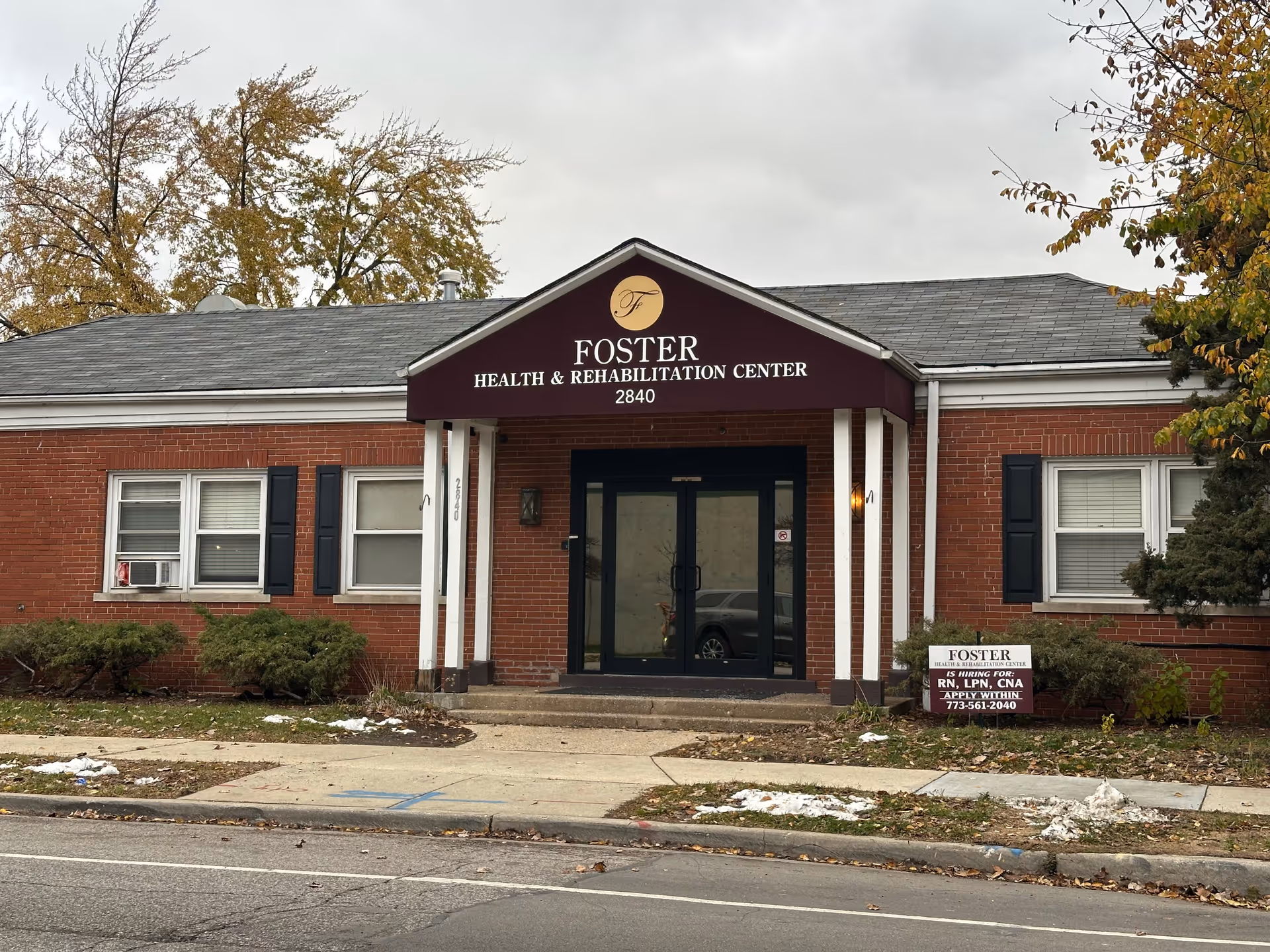 Front exterior view of Foster Health & Rehabilitation Center, a single-story brick building with a maroon awning displaying the facility name and address 2840. There are bushes and trees around the building, a sidewalk in front, and a sign near the entrance advertising job openings for RN, LPN, and CNA positions with a contact phone number.