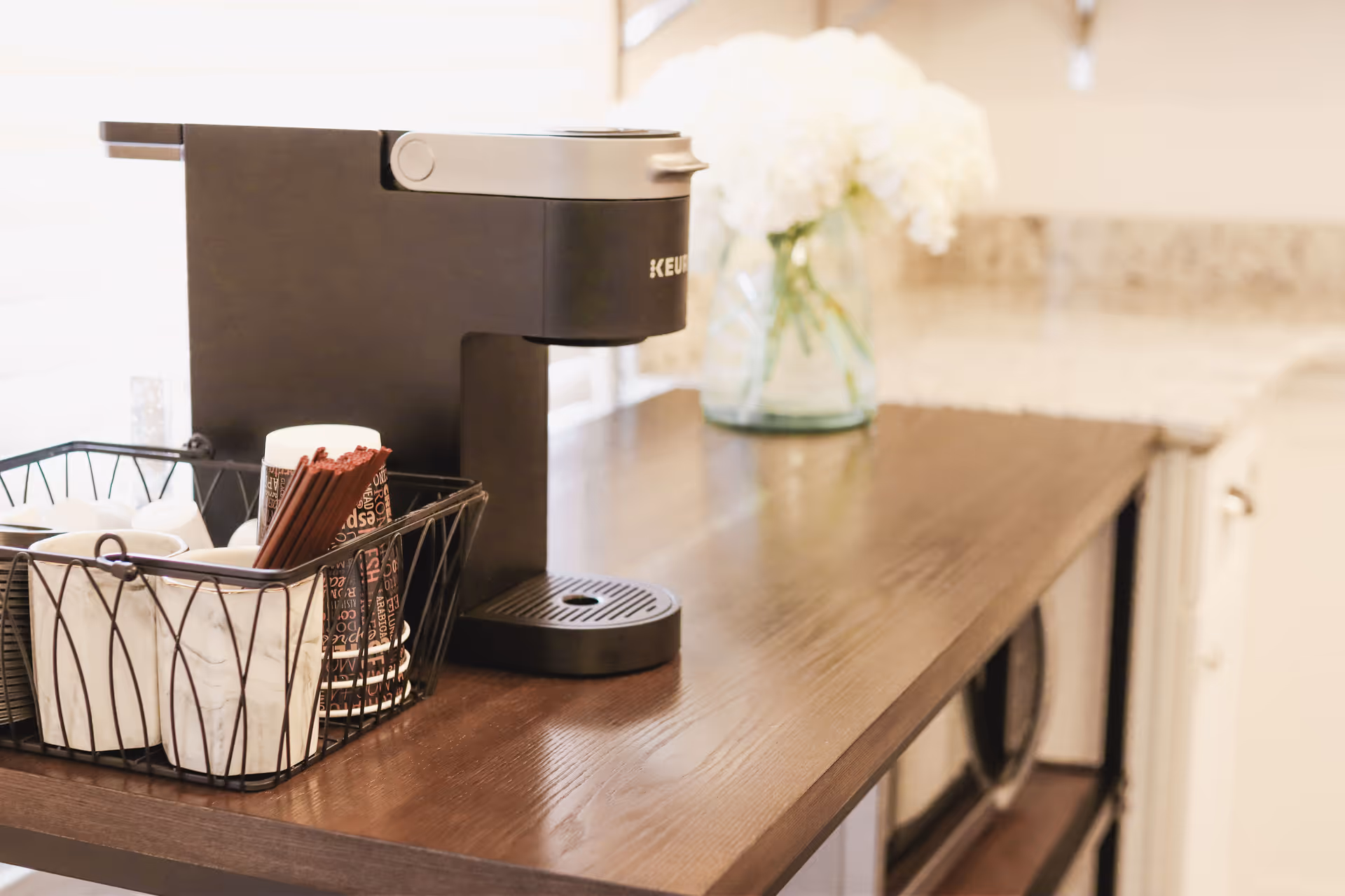 Black coffee machine on a wooden counter with cups and sugar packets, flowers in background.