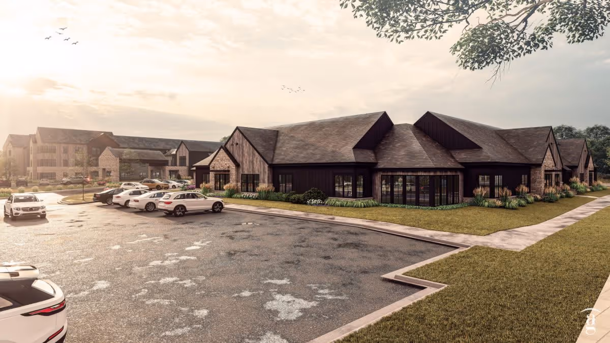 Exterior view of a senior living facility named FeatherStone at Hickory Hill during sunset, showing a large parking lot with several parked cars, a modern building with multiple peaked roofs, stone and wood siding, surrounded by landscaped grass and trees.