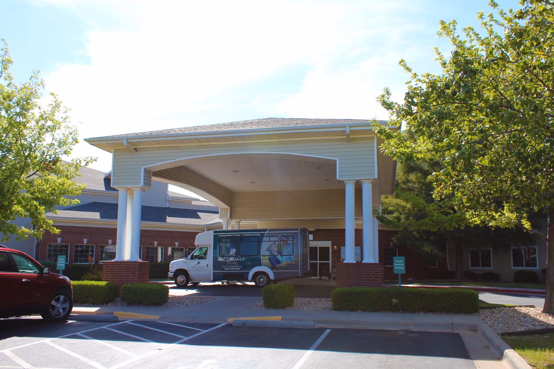 Front entrance of Lyndale Abilene Senior Living facility with a covered drop-off area supported by white columns. A shuttle bus is parked under the covered area, and there are trees and bushes around the parking lot.