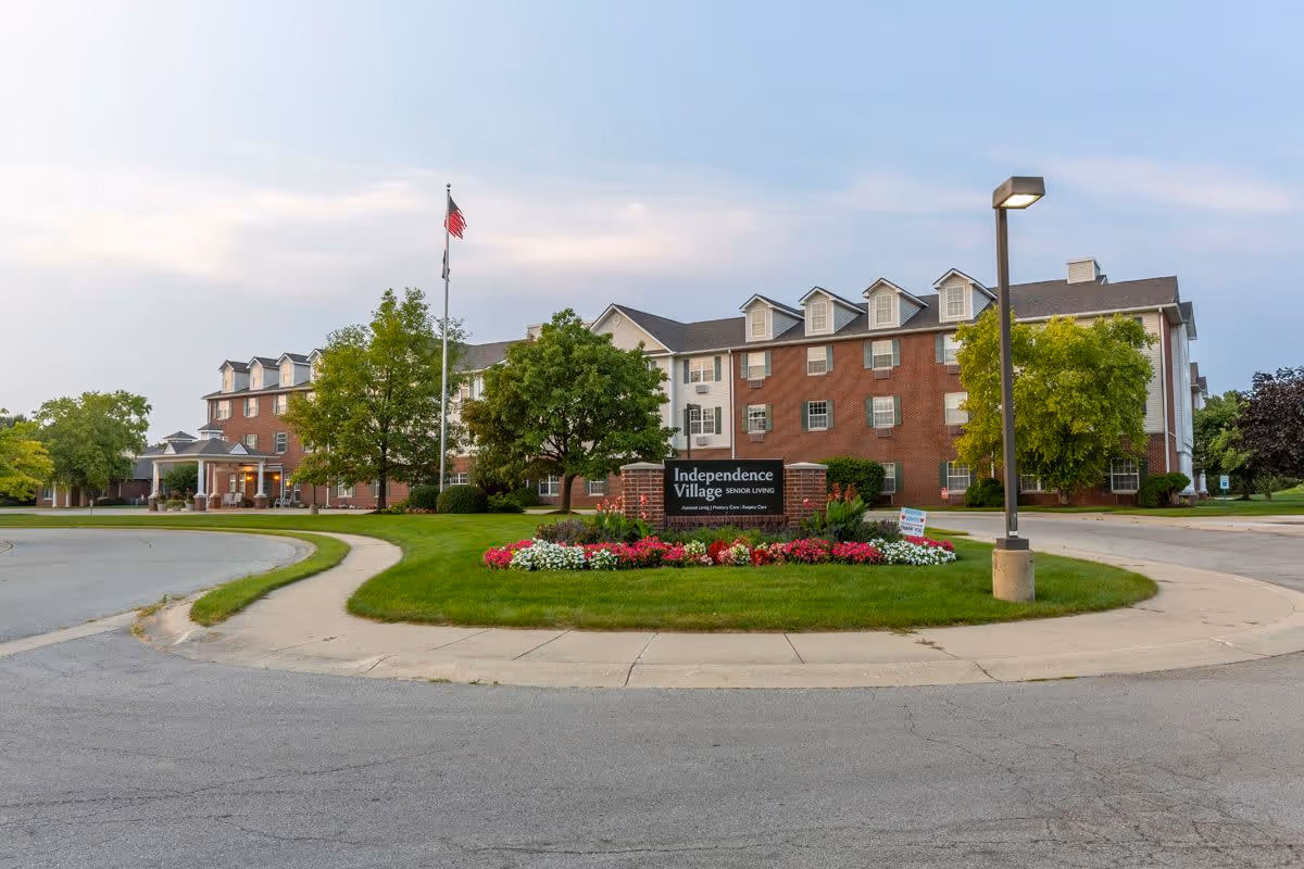 Exterior view of Independence Village of Fishers South, a senior living facility with a large brick and white building, surrounded by green trees and a well-maintained lawn with colorful flowers. An American flag is flying on a flagpole near the entrance.