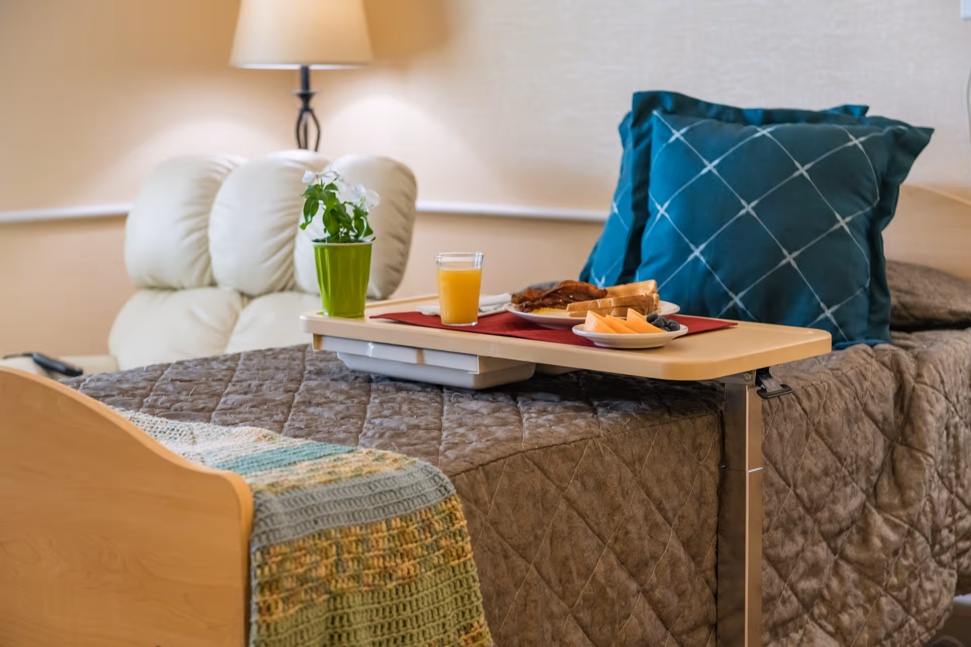 A bed with a breakfast tray, plant, and pillows sits in a cozy senior living bedroom.