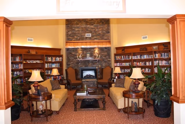 Cozy library room with two yellow sofas facing each other, a wooden coffee table in the center, two side tables with lamps, and a stone fireplace with two armchairs on either side. Bookshelves filled with books line the back wall, and there are potted plants on either side of the seating area.